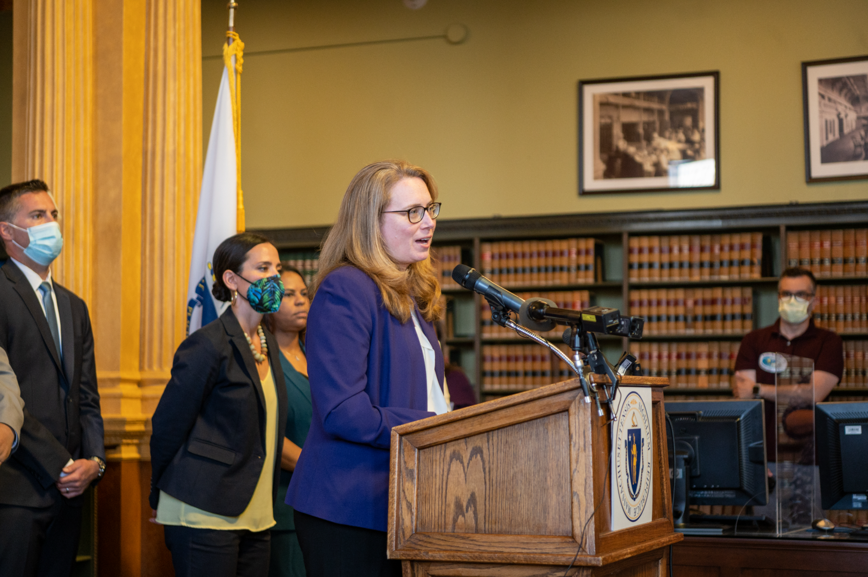 Christine Barber speaking at a podium at the State House, flanked by colleagues and the Massachusetts flag
